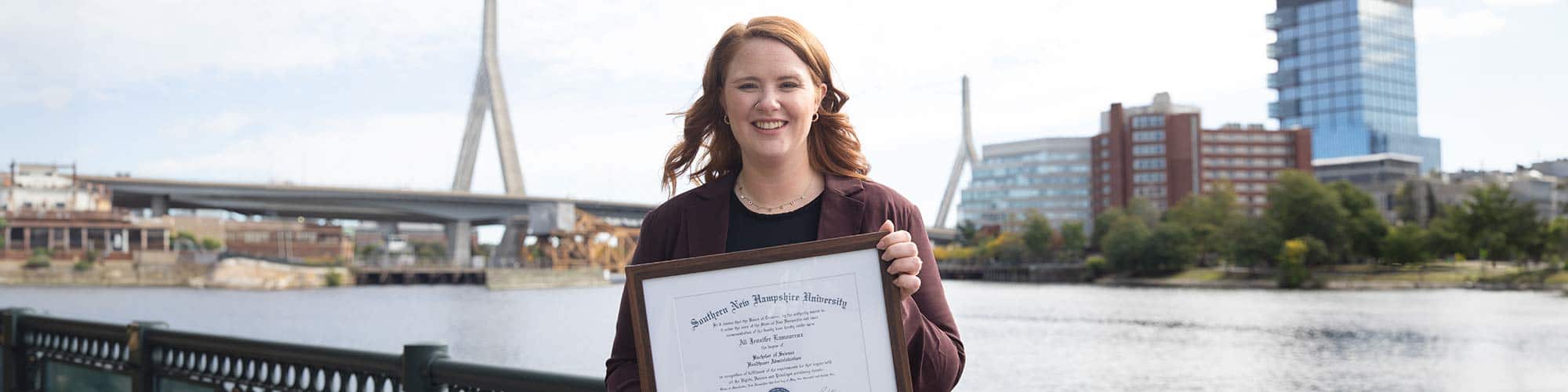 A young, smiling female student holds her framed SNHU diploma in front of a body of water