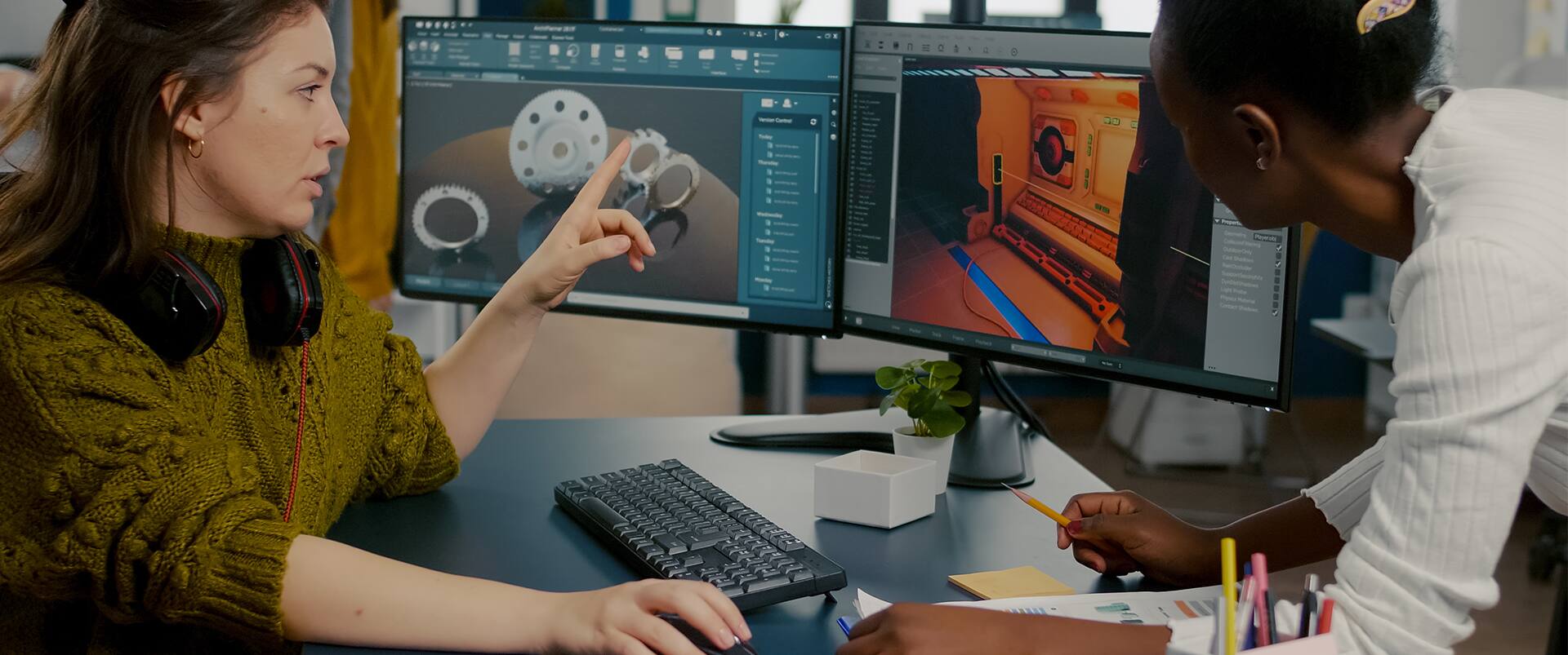 Two professionals collaborate at a workstation with dual monitors displaying 3D modeling and game design software, one pointing at the screen.