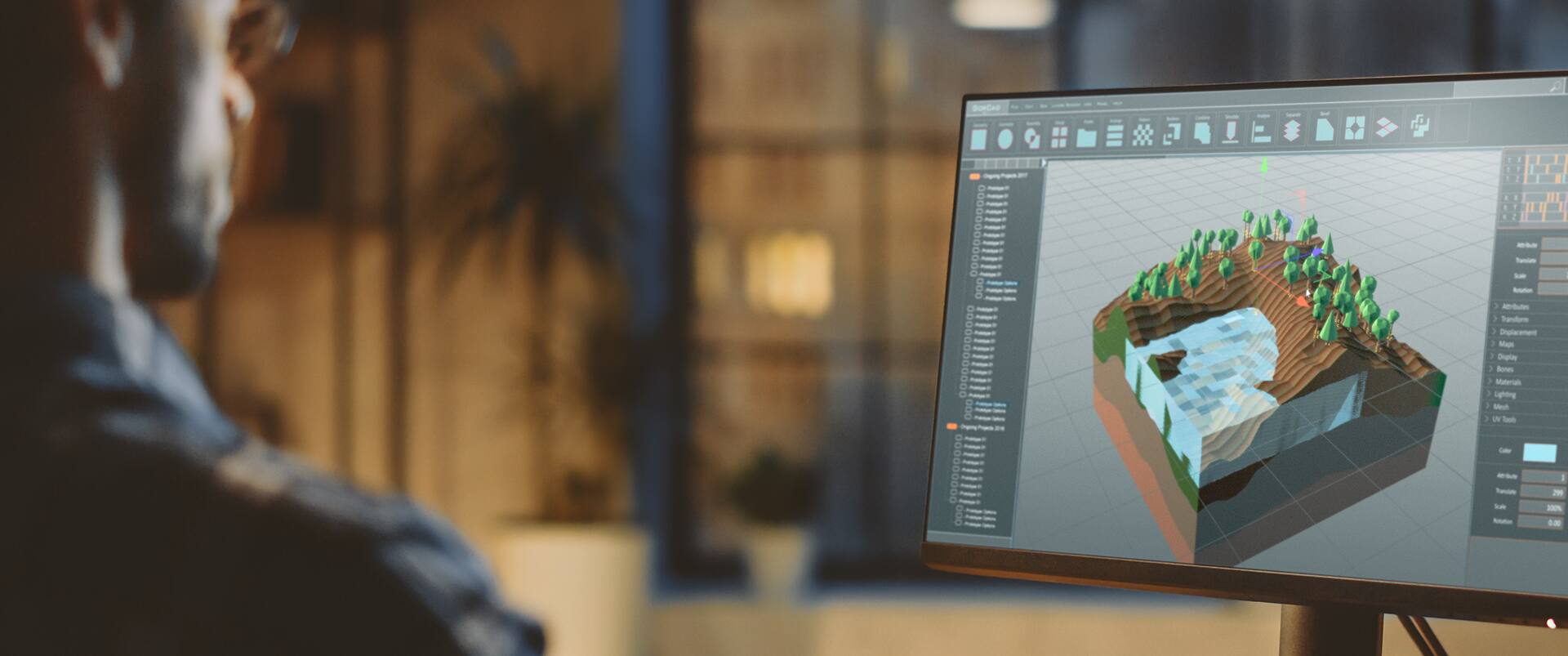 A game designer works at a monitor displaying a low-poly 3D landscape environment with trees and water, built in 3D modeling software.