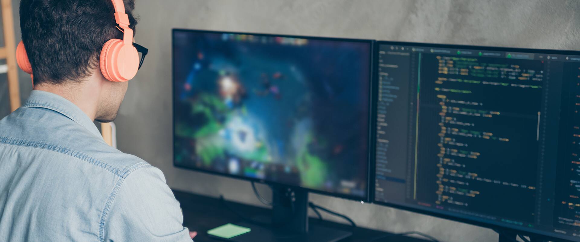 A game programmer wearing headphones works at dual monitors showing a fantasy video game and a code editor with colorful syntax highlighting.