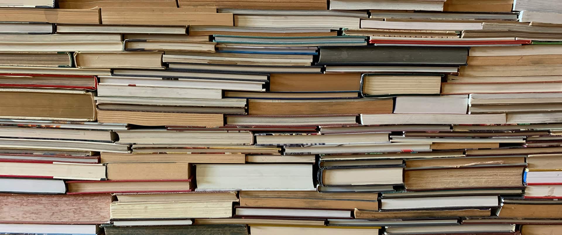 Close-up of hardcover books stacked horizontally, page edges facing out, forming a dense textured wall of tan, cream, and beige tones with occasional colorful spines.