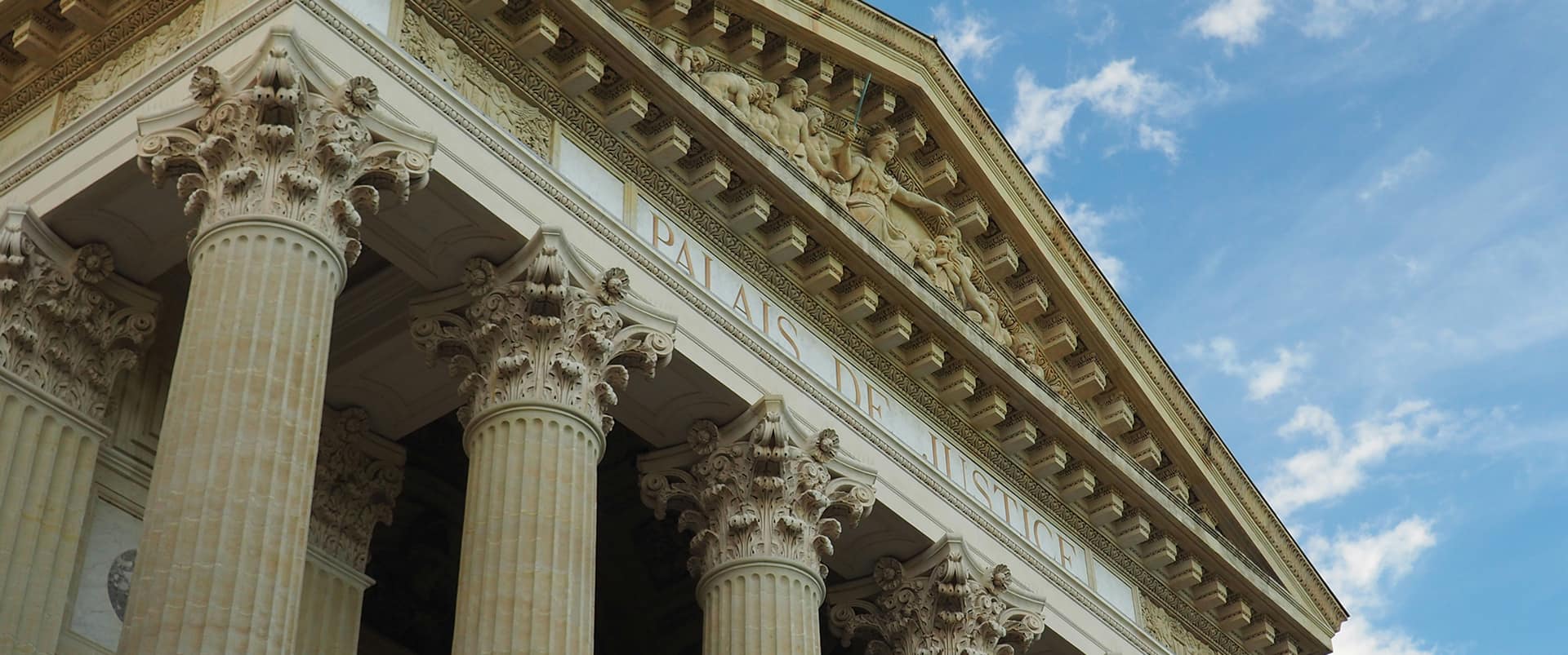 Upward view of a "Palais de Justice" facade with ornate Corinthian columns, a carved pediment depicting robed figures and Lady Justice, set against a blue sky with light clouds.