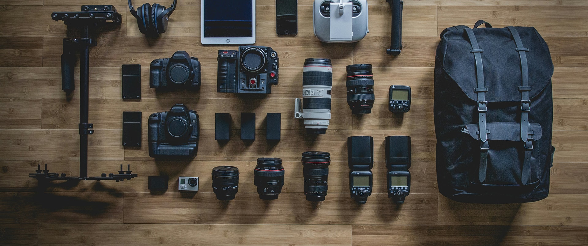 Flat lay of professional camera gear on a wood floor: two DSLR bodies, a cinema camera, GoPro, several lenses, flashes, a stabilizer rig, drone remote, tablet, phone, headphones, and a black backpack.